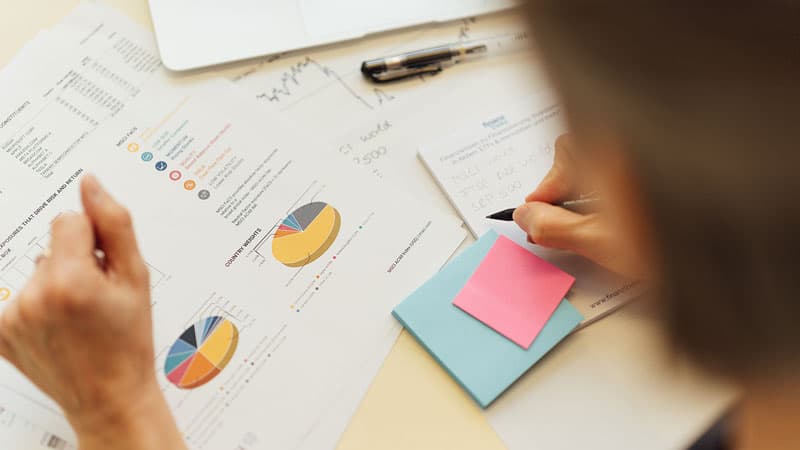 Person, die gedruckte Diagramme und Grafiken mit bunten Tortendiagrammen betrachtet, Notizen auf Papier neben Haftnotizen und Finanzdokumenten schreibt, mit einem Stift und einem teilweise sichtbaren offenen Laptop.