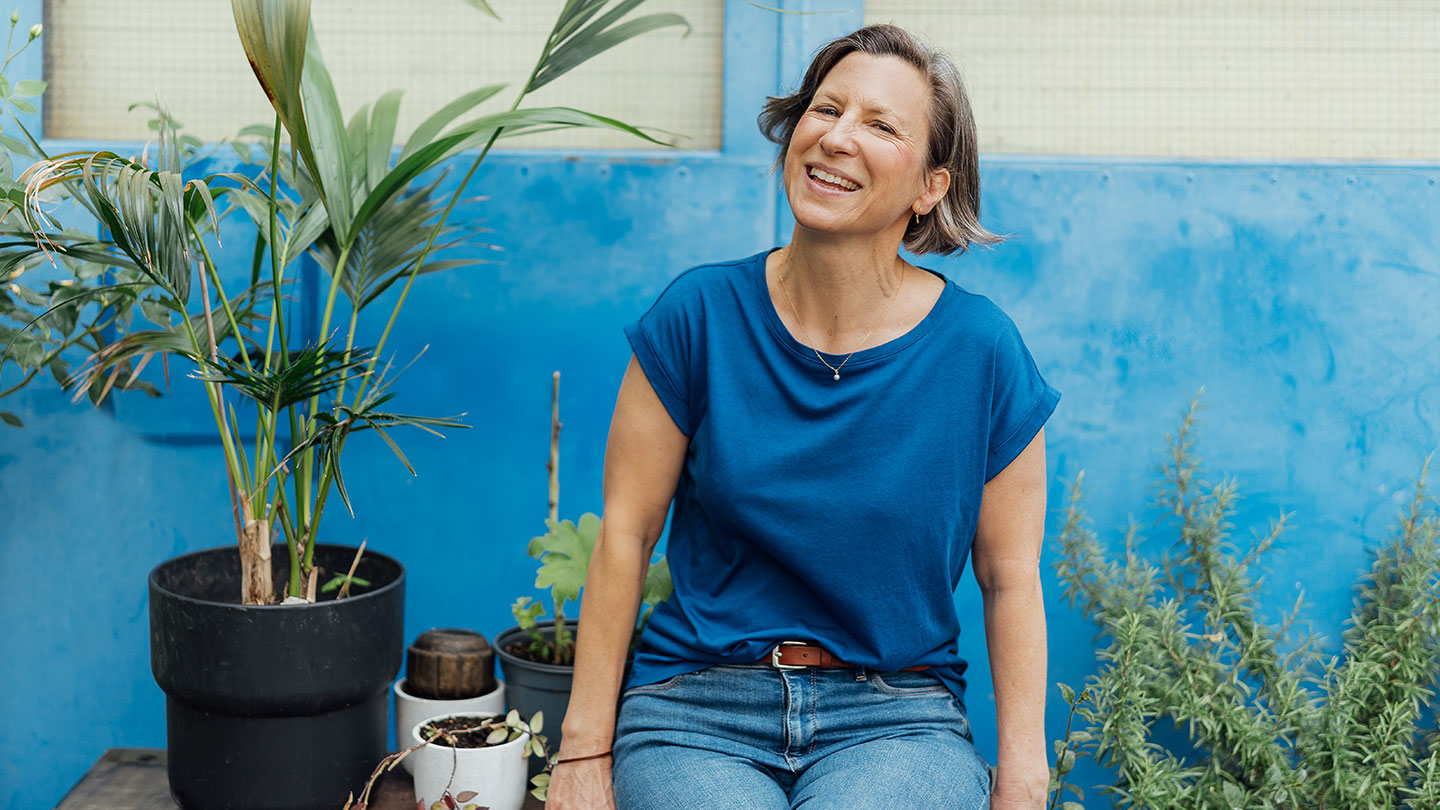 Eine Frau in blauem T-Shirt und Jeans sitzt lächelnd neben Topfpflanzen an einer hellblauen Wand in einem sonnendurchfluteten Innengarten.