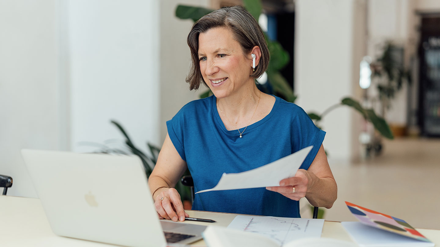 Eine Frau mit kurzen Haaren und blauem Hemd sitzt an einem Schreibtisch und lächelt in ihren Laptop. Sie hält ein Blatt Papier in der Hand und trägt kabellose Ohrstöpsel. Auf dem Tisch in einem hellen Büroraum liegen Dokumente und ein Stift.
