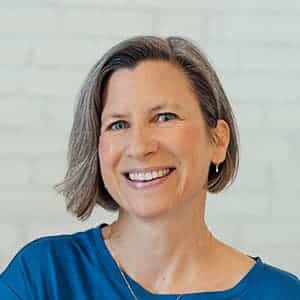 Eine Frau mit kurzem, graubraunem Haar lächelt in die Kamera. Sie trägt ein blaues Oberteil und einen kleinen Creolenohrring. Der Hintergrund ist eine weiße Backsteinmauer.