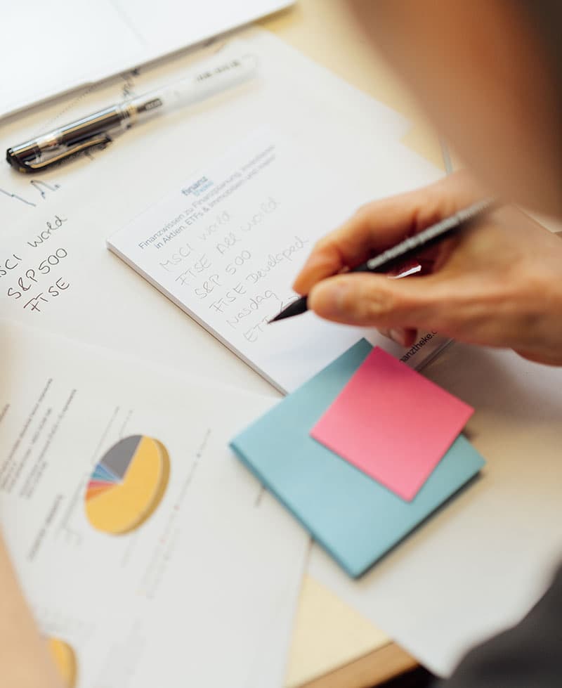 Eine Person schreibt Notizen auf einem Block neben bunten Haftnotizen und Diagrammen mit Kuchendiagrammen auf einem Schreibtisch, neben einem Stift und einem Blatt mit handgeschriebenen Aktiensymbolen.