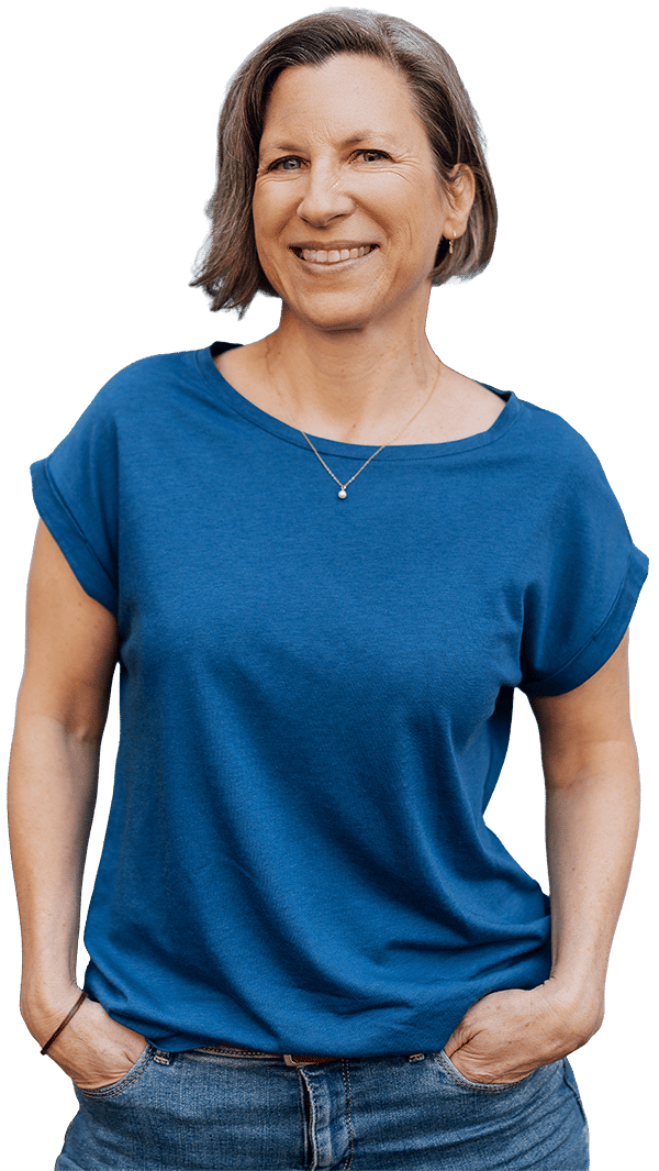 Eine lächelnde Frau mit kurzen braunen Haaren, die ein blaues kurzärmeliges Hemd und Jeans trägt, steht mit den Händen in den Taschen vor einem schlichten weißen Hintergrund.