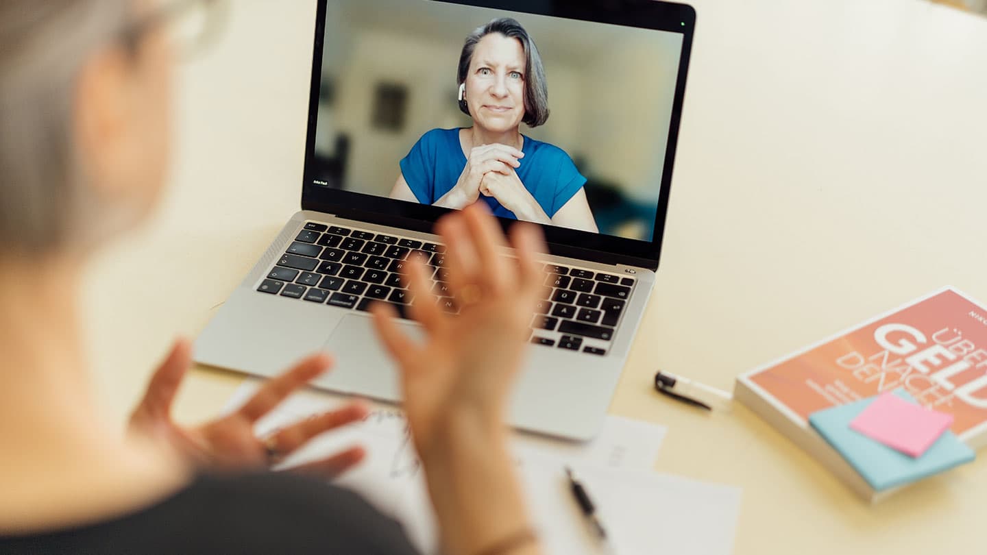 Eine Person nimmt an einem Videogespräch auf einem Laptop teil und gestikuliert mit ihren Händen. Eine andere Frau erscheint auf dem Bildschirm des Laptops und hört aufmerksam zu. Auf dem Schreibtisch daneben liegen ein Buch, ein Stift und Papiere.