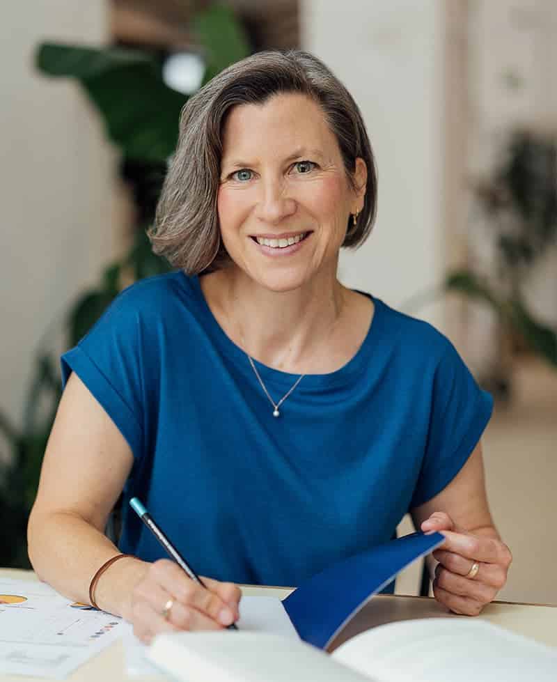 Eine lächelnde Frau mit kurzen grauen Haaren und einem blauen Hemd sitzt an einem Tisch, hält einen Stift und ein Buch in der Hand und scheint zu schreiben oder Notizen zu machen. Im Hintergrund sind Pflanzen und weiches Licht zu sehen.