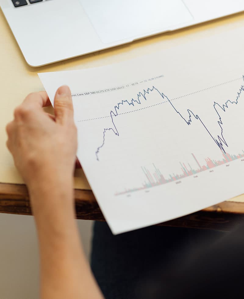 Eine Person hält ein gedrucktes Blatt mit einem schwankenden Linien- und Balkendiagramm in der Hand und wertet Daten aus. Ein Teil eines Laptops ist auf dem Schreibtisch über dem Blatt zu sehen.