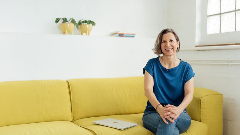 Eine Frau sitzt auf einer gelben Couch und lächelt mit verschränkten Händen. Sie trägt ein blaues Hemd und Jeans. Auf der Couch neben ihr steht ein zugeklappter Laptop. Hinter ihr befinden sich zwei Topfpflanzen und ein Stapel Bücher auf einem weißen Regal.